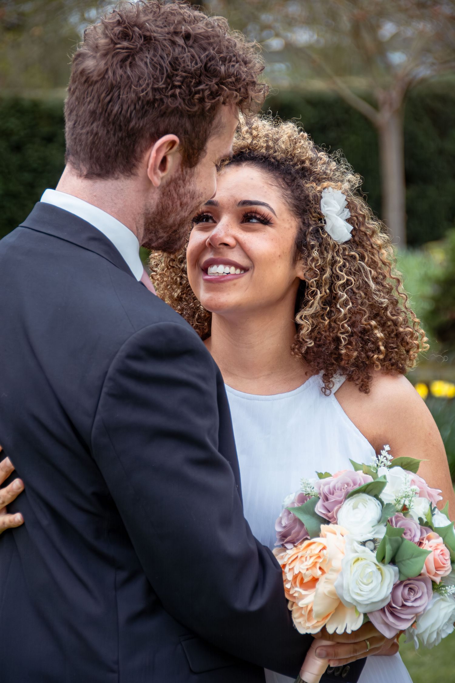 A smiling curly-haired bride holding a pastel bouquet looks up as her groom kisses her cheek outdoors.