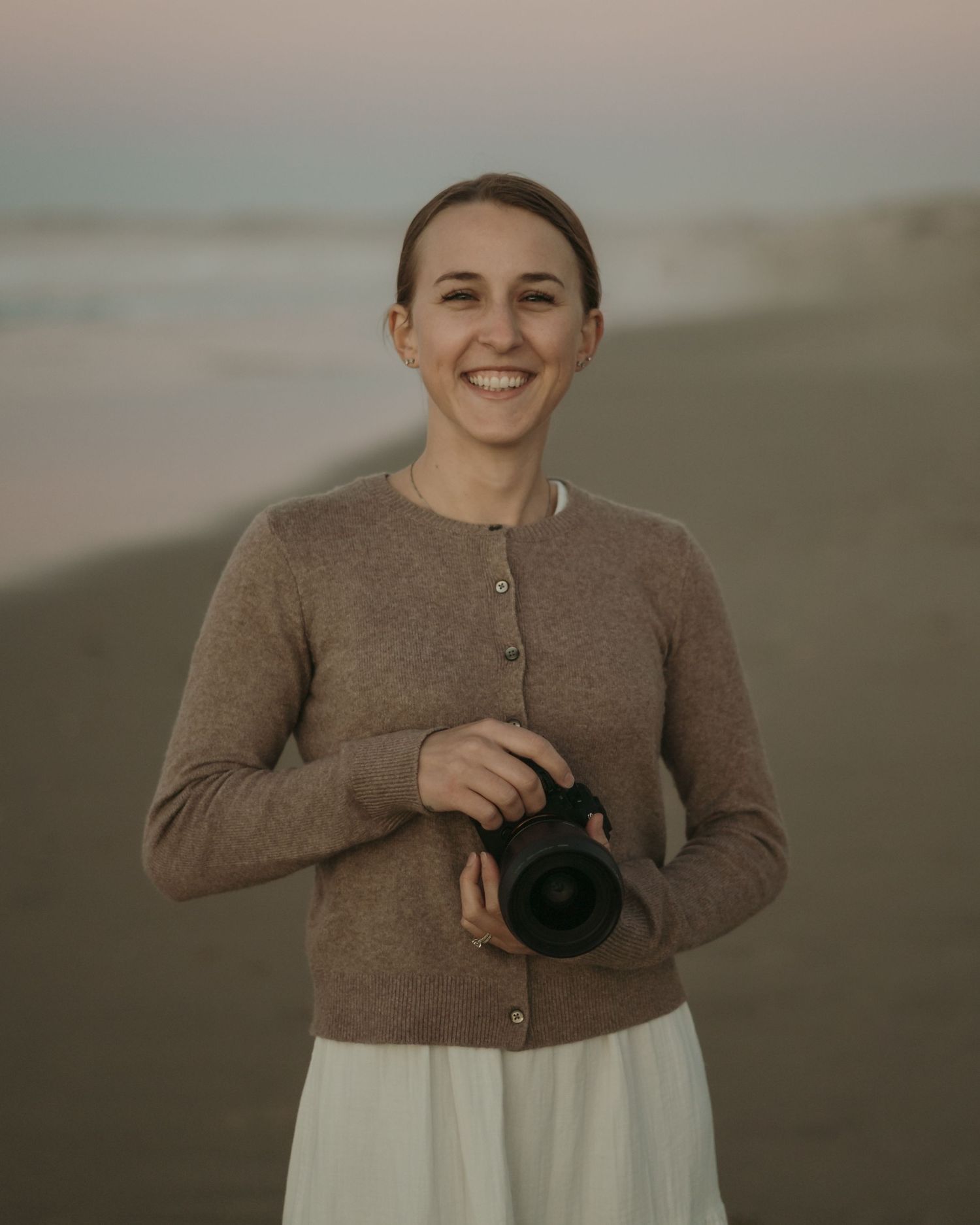 Person wearing black sweater and beige wide-leg pants poses outdoors near wooden fence in autumn lighting.