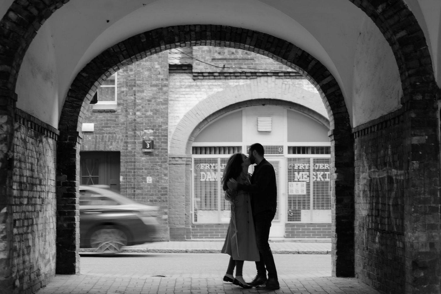 A silhouetted couple embraces beneath an arched passageway while a car blurs past in the black and white scene.