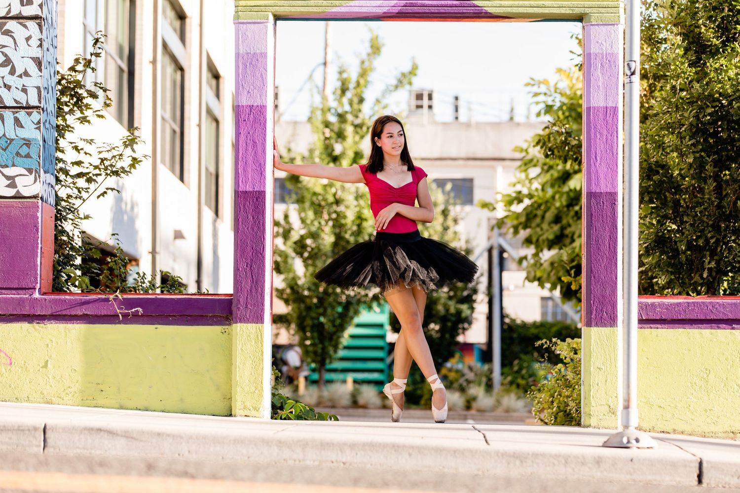 A dancer in a black tutu and red top poses elegantly against a colorful outdoor urban backdrop with pink framing.