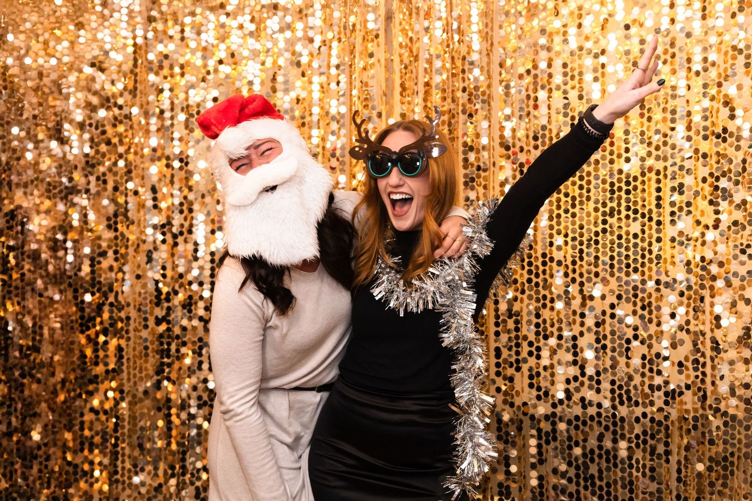 Two people pose energetically in front of a gold sequin backdrop with holiday props at a festive photo booth.
