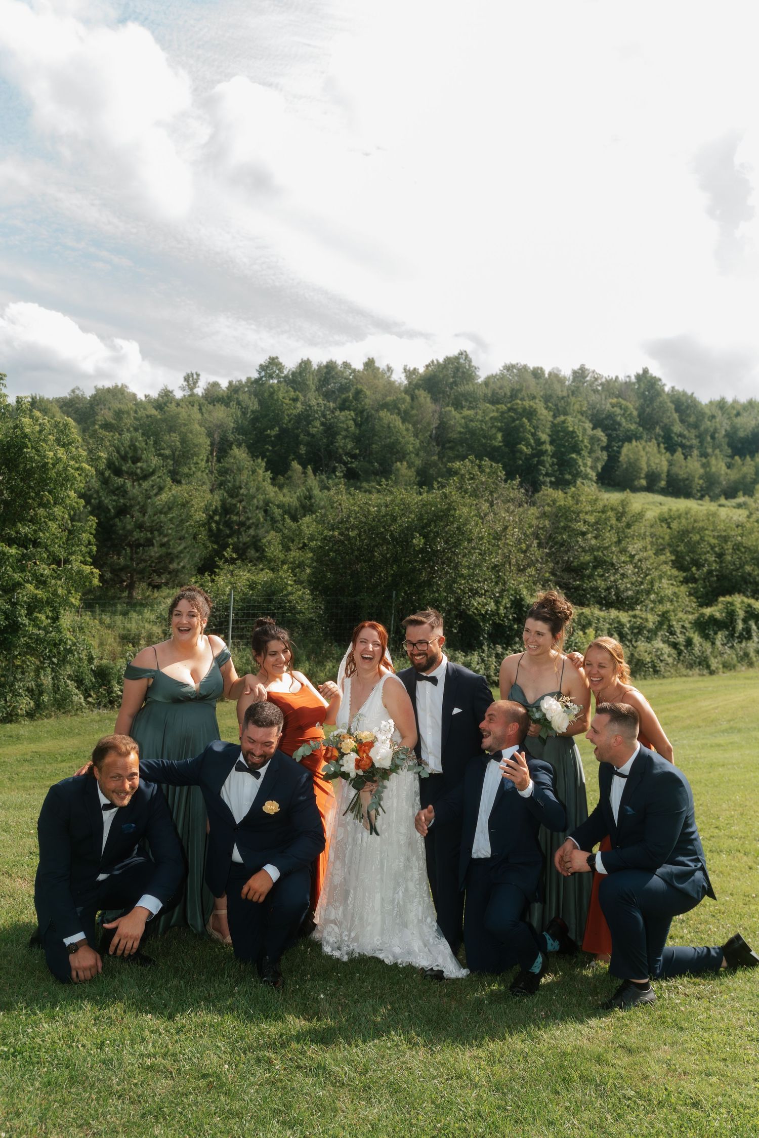 Wedding party poses on green lawn with lush forest backdrop, bride in lace gown with colorful bouquet.