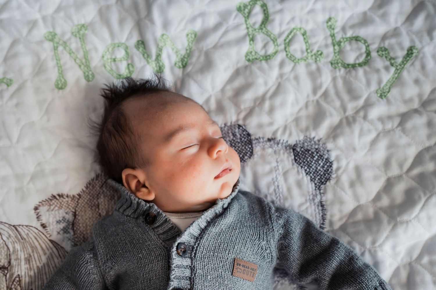Adorable newborn swaddled in gray knit blankets sleeps peacefully on white ruffled bedding.