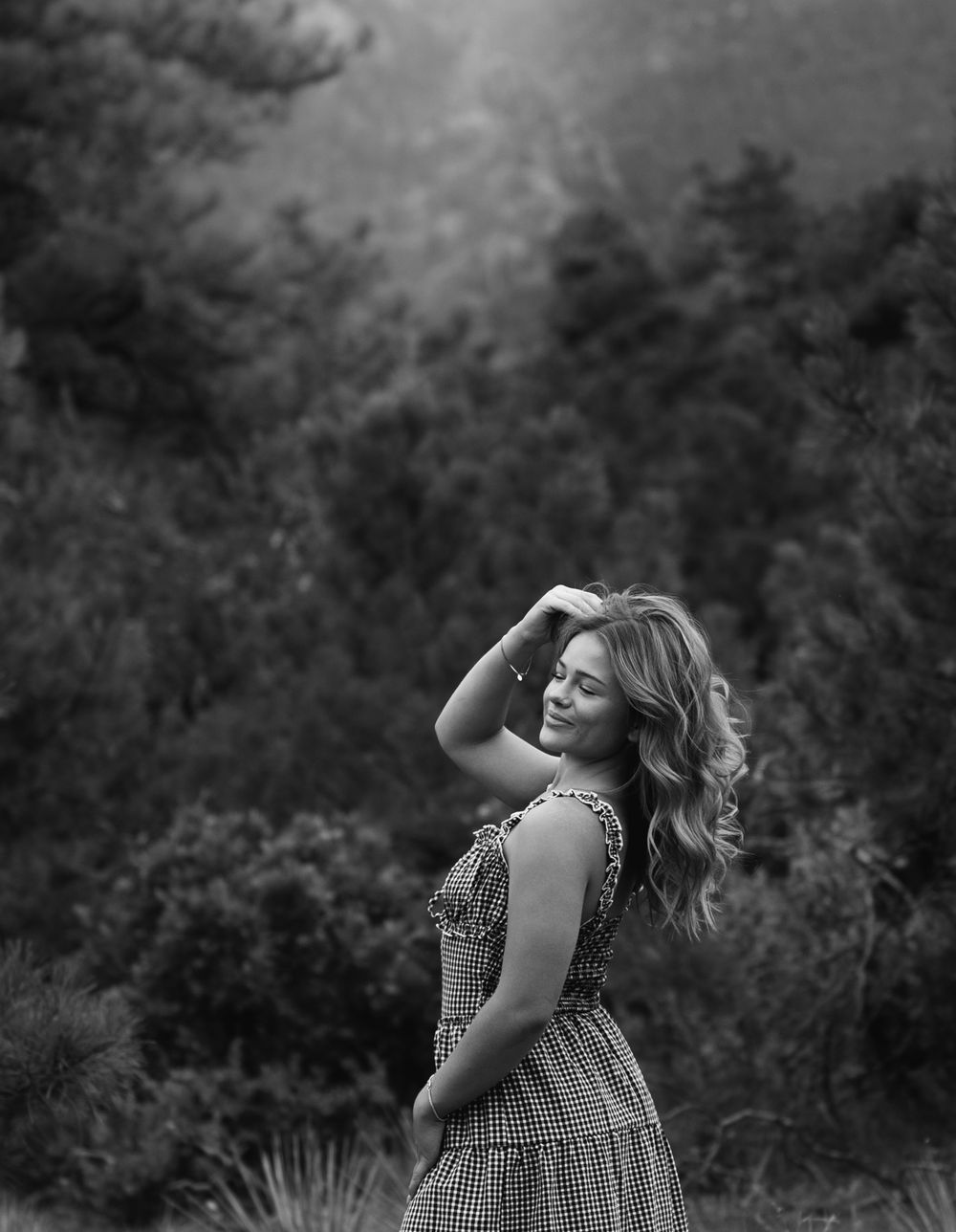A person in a sleeveless gingham dress walks through a lush garden setting in black and white photography.