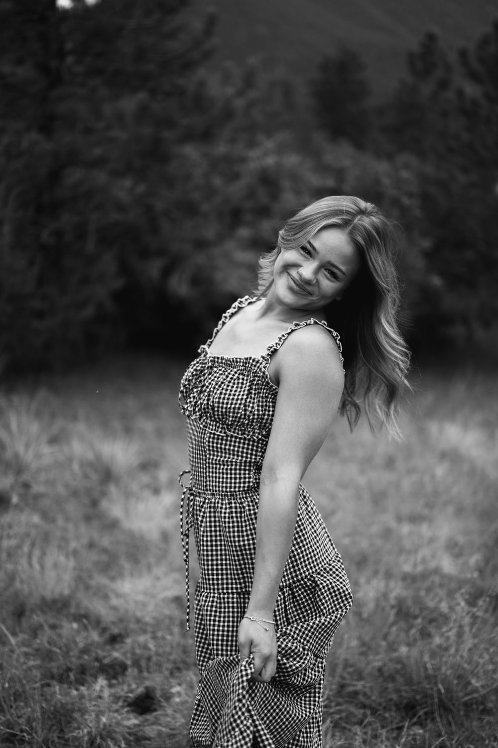 Black and white portrait of a woman in a gingham dress posing expressively in a grassy outdoor setting.