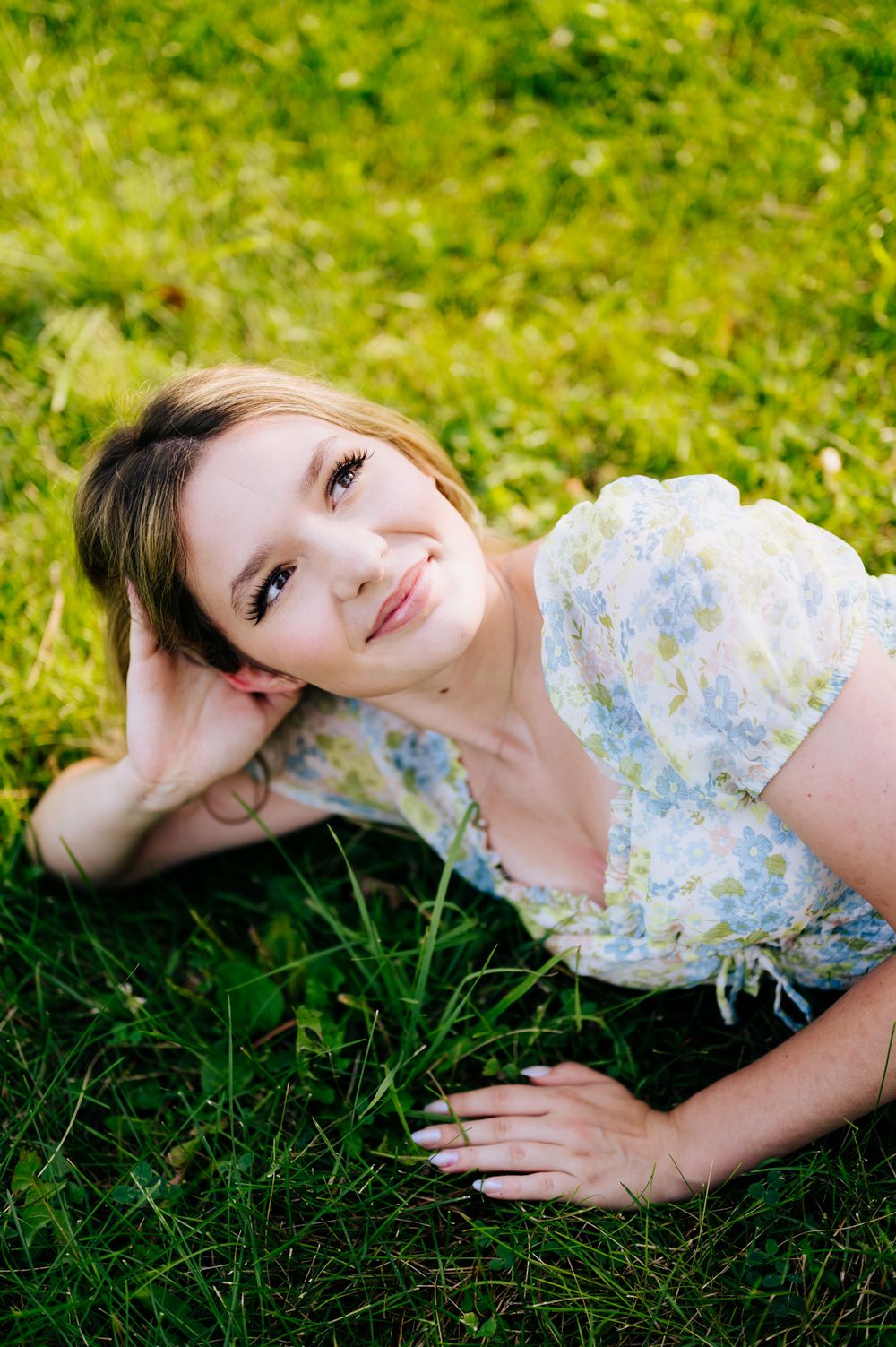 A person in a floral dress lies peacefully on vibrant green grass in a sunny outdoor setting.