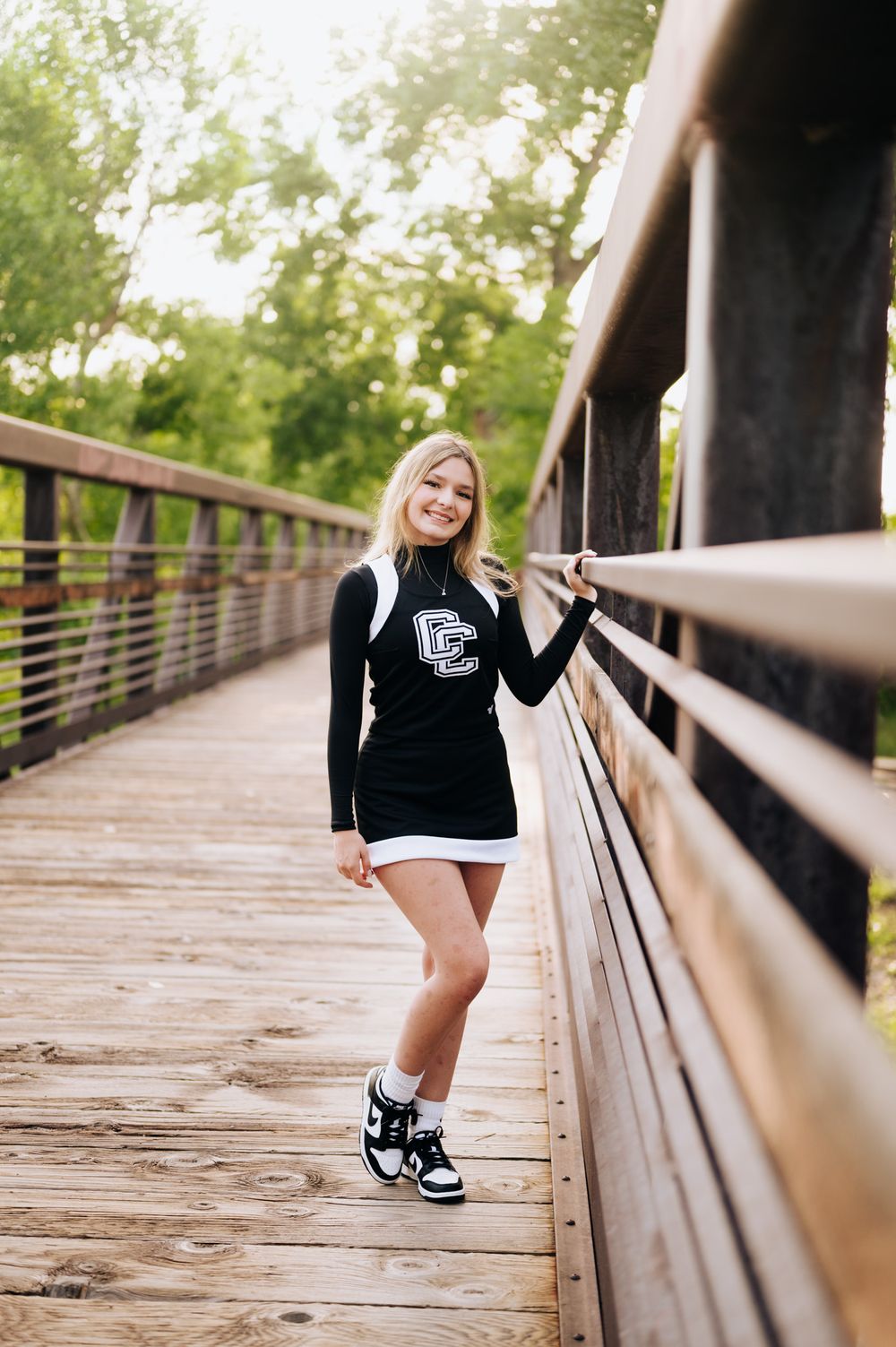 A cheerleader in black and white uniform poses on a wooden bridge walkway in a natural outdoor setting.