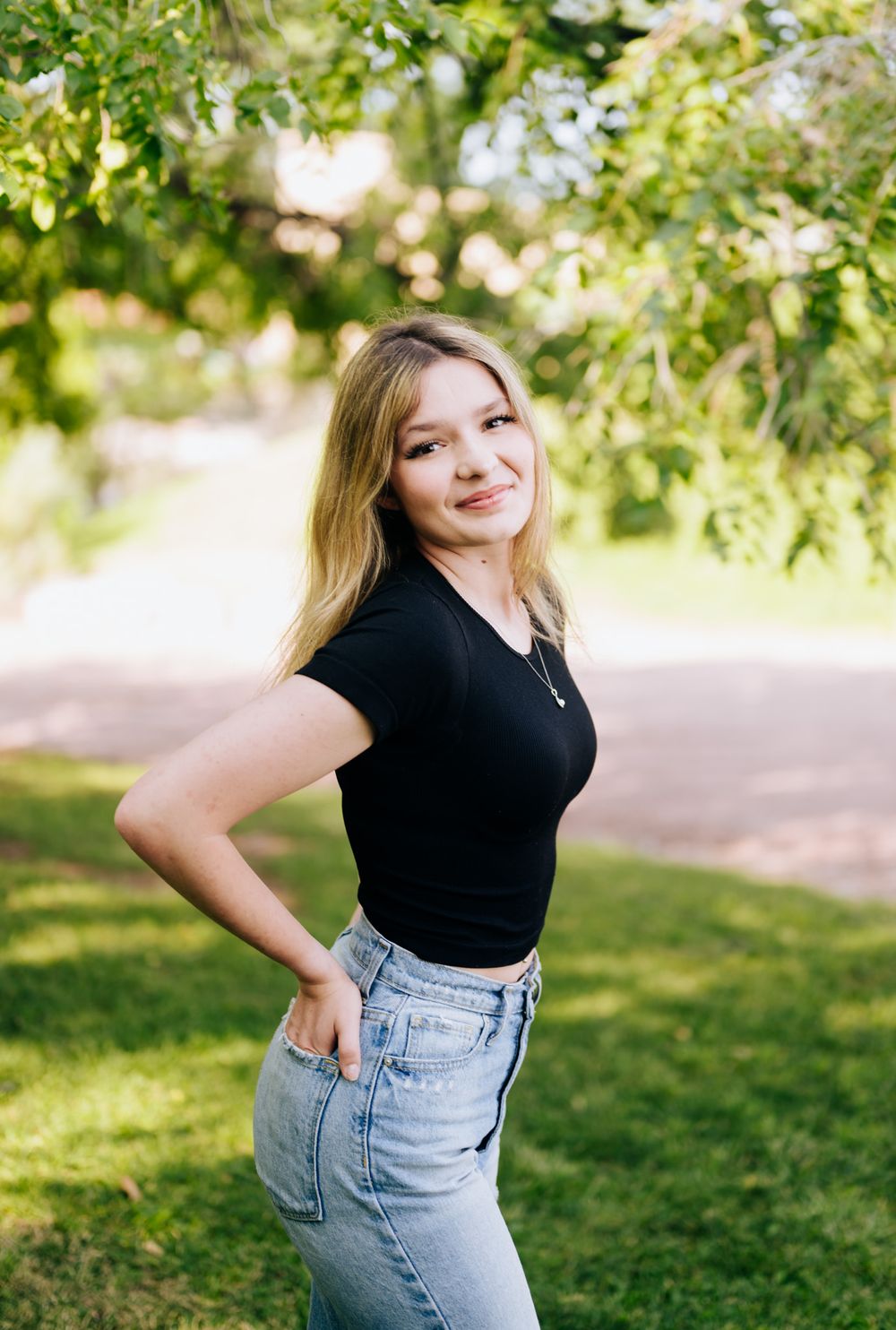 Person in a black top and high-waisted jeans poses playfully in a sunny park setting with green foliage in background.