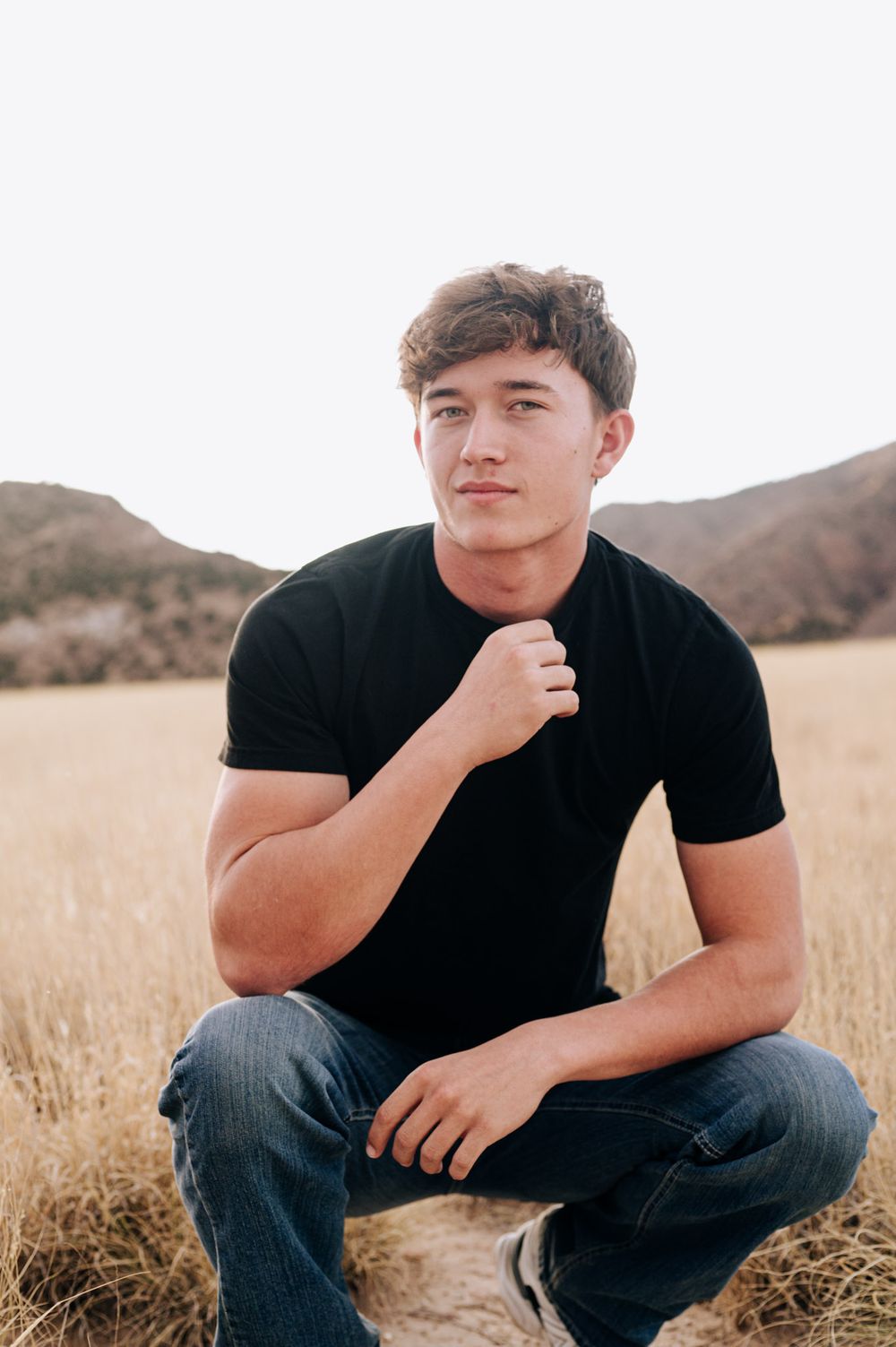Young person in black t-shirt sits thoughtfully in golden grass field with mountains in background.
