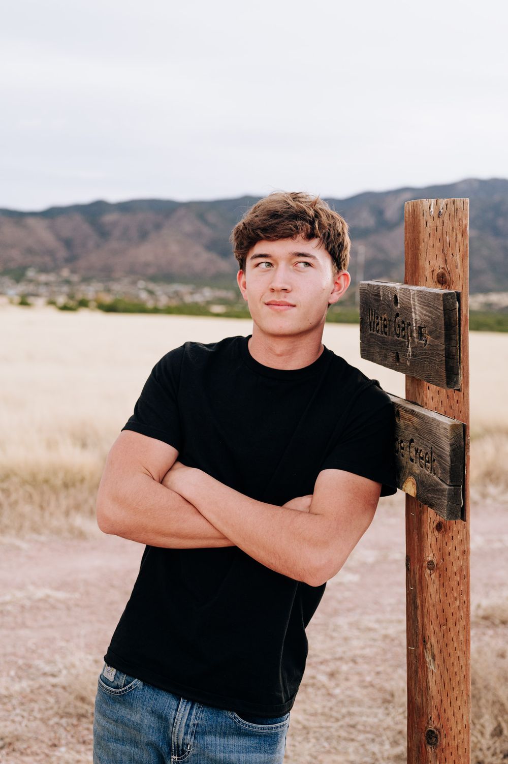 A person in a black t-shirt and jeans poses casually against a wooden fence post in a desert landscape.