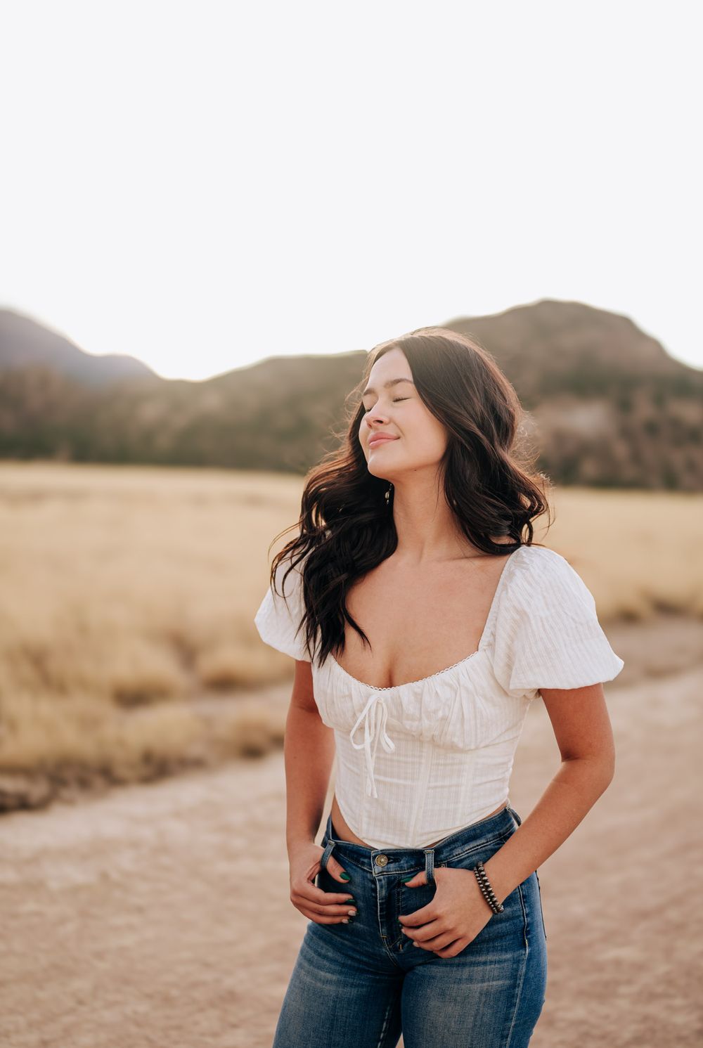 A person in a white peasant top and jeans poses playfully in a desert landscape during golden hour.