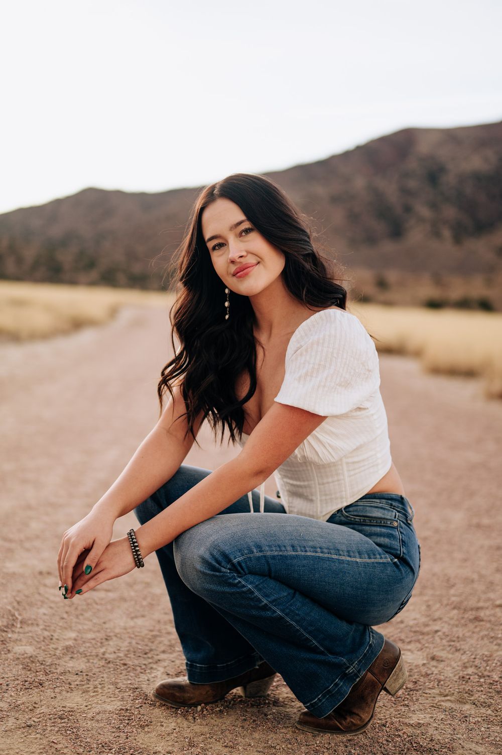 A person in a white top and jeans crouches in an arid field with mountains visible in the background.