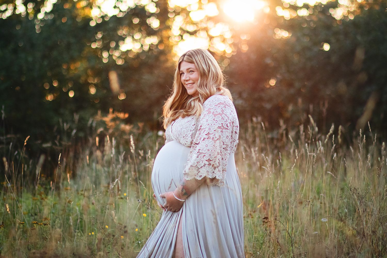 Smiling pregnant woman in lace maternity gown holds belly in golden sunset meadow.
