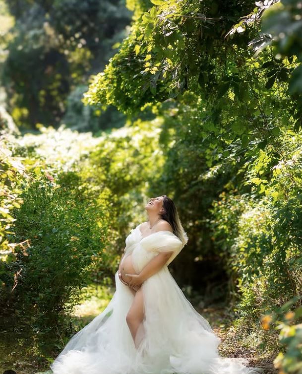 A figure in a flowing white wedding dress poses gracefully among lush green garden foliage in soft sunlight.