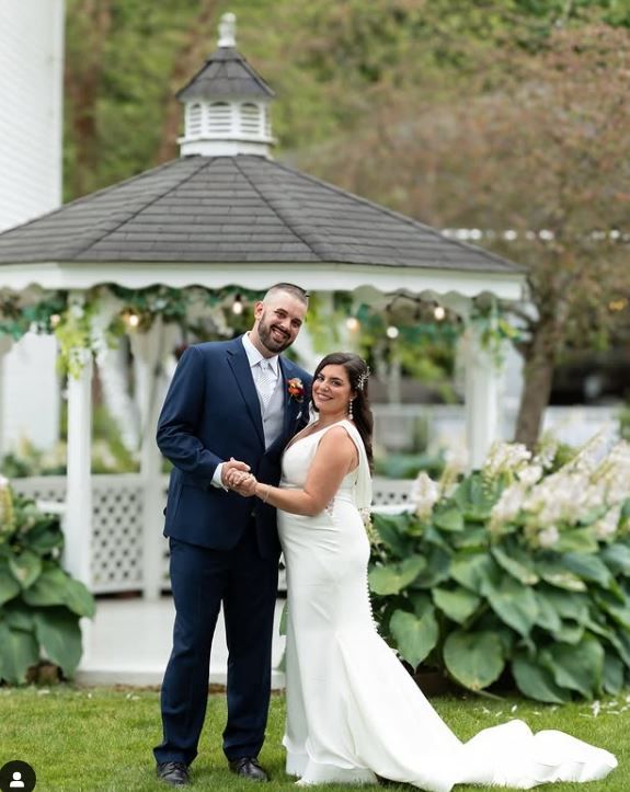 A couple shares a romantic moment in front of a white gazebo surrounded by lush garden landscaping.