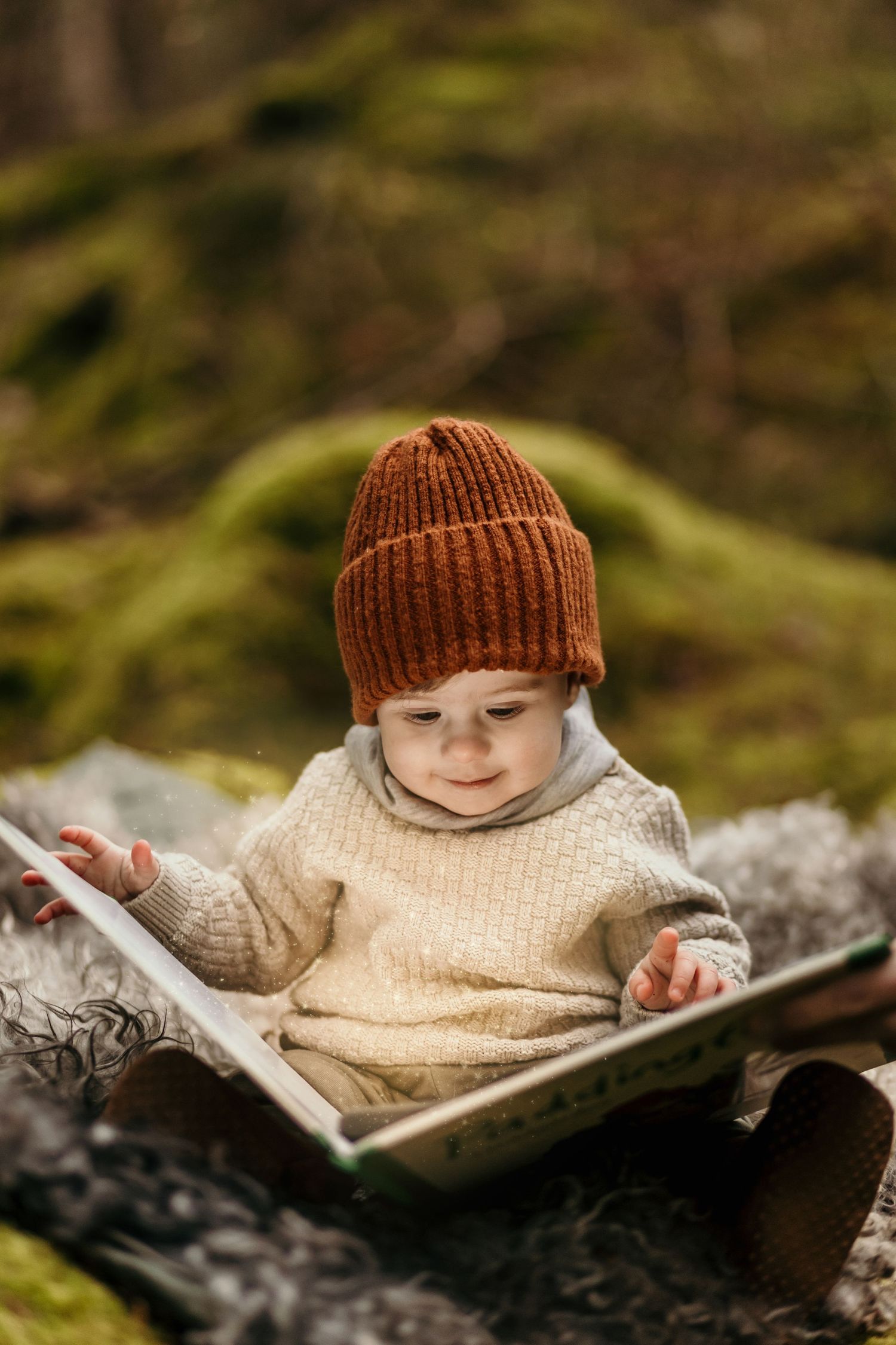 Small figure in brown knit hat and cream sweater reading a book while sitting on mossy ground in nature setting.