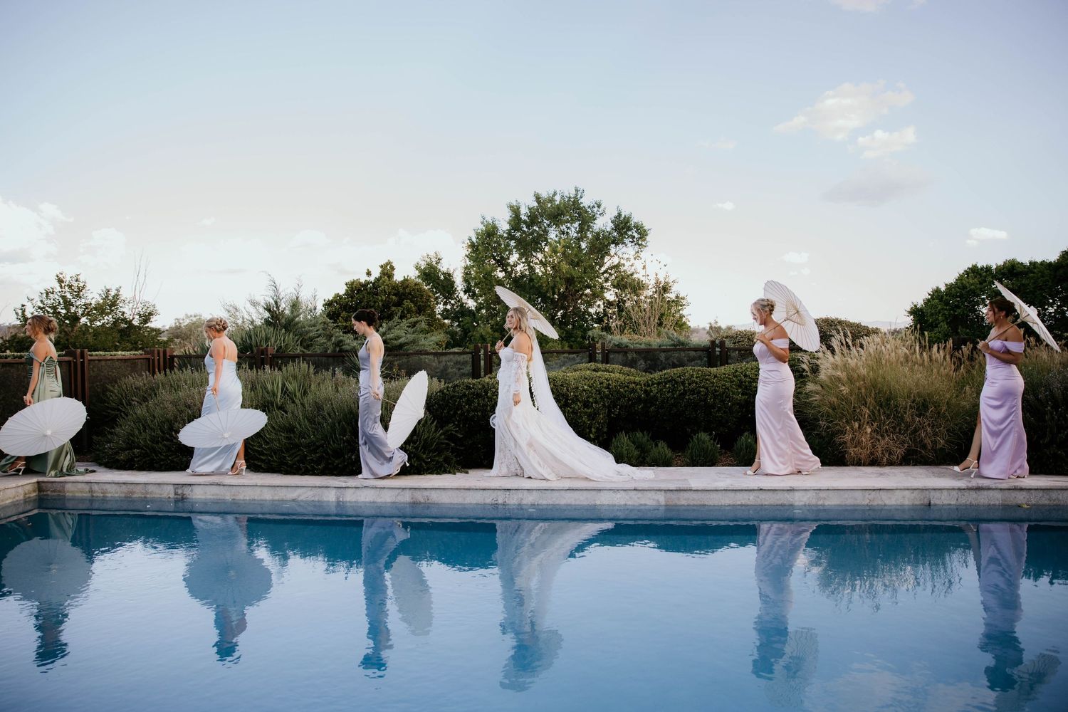 Row of bridesmaids in light-colored dresses walking along a poolside at sunset, creating reflections in the blue water.