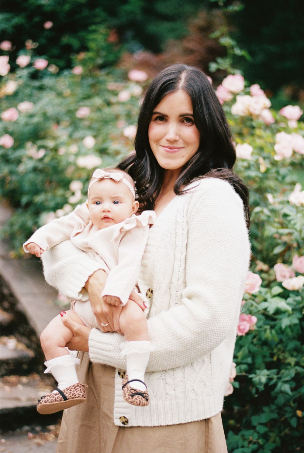 Baby in cream-colored outfit being held while standing in a garden with blooming pink roses in background.