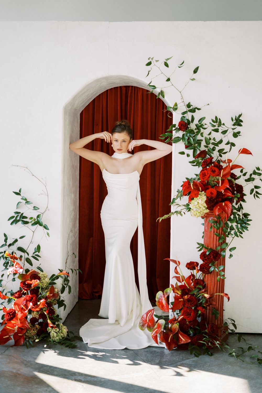 A white wedding gown displayed in an archway adorned with vibrant red floral arrangements and greenery.