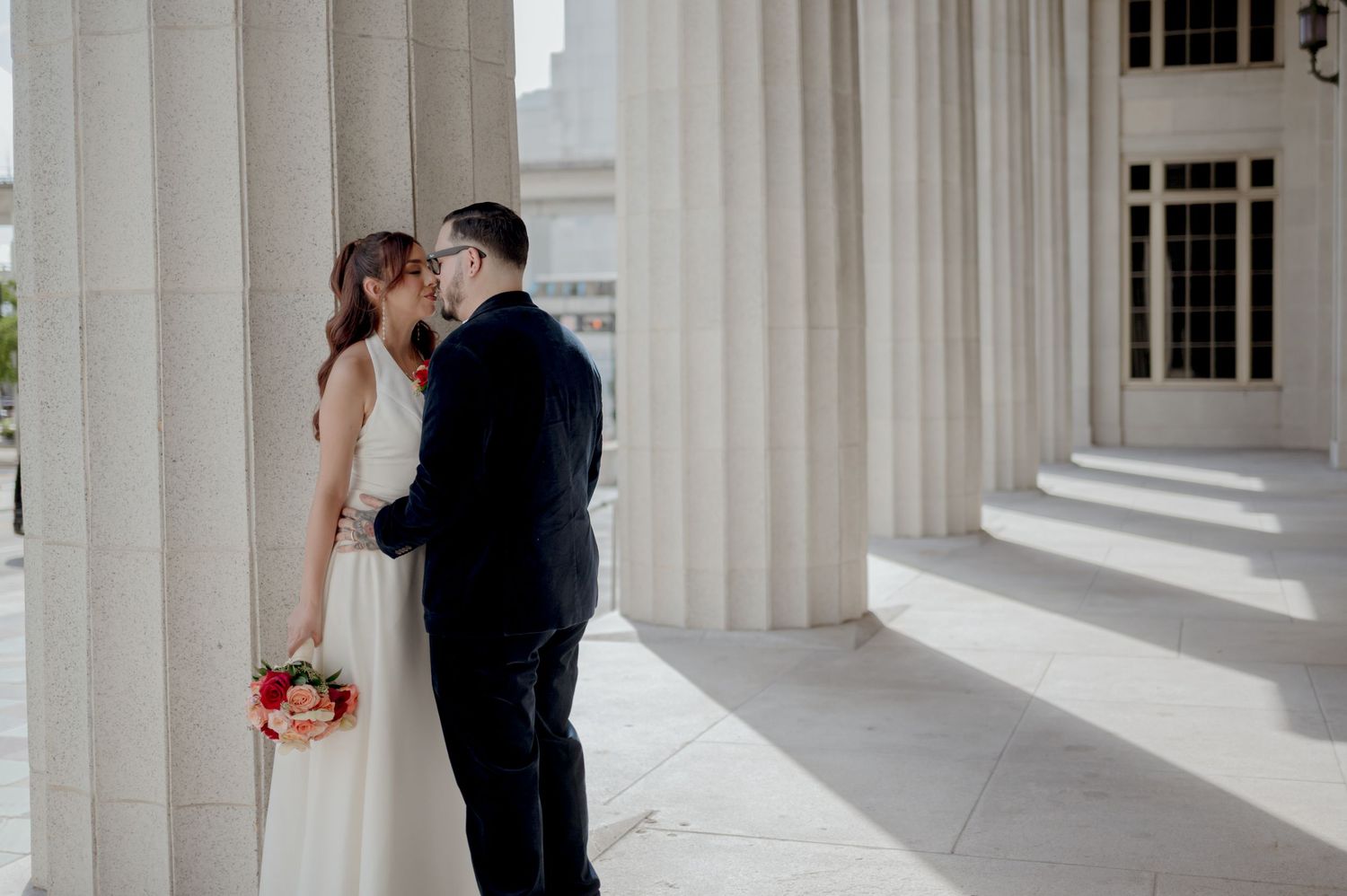 A bride and groom share a romantic kiss under elegant white columns of a classic building.