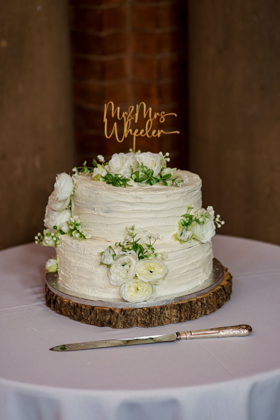 Elegant white tiered wedding cake decorated with fresh flowers sits on wooden slice platter.