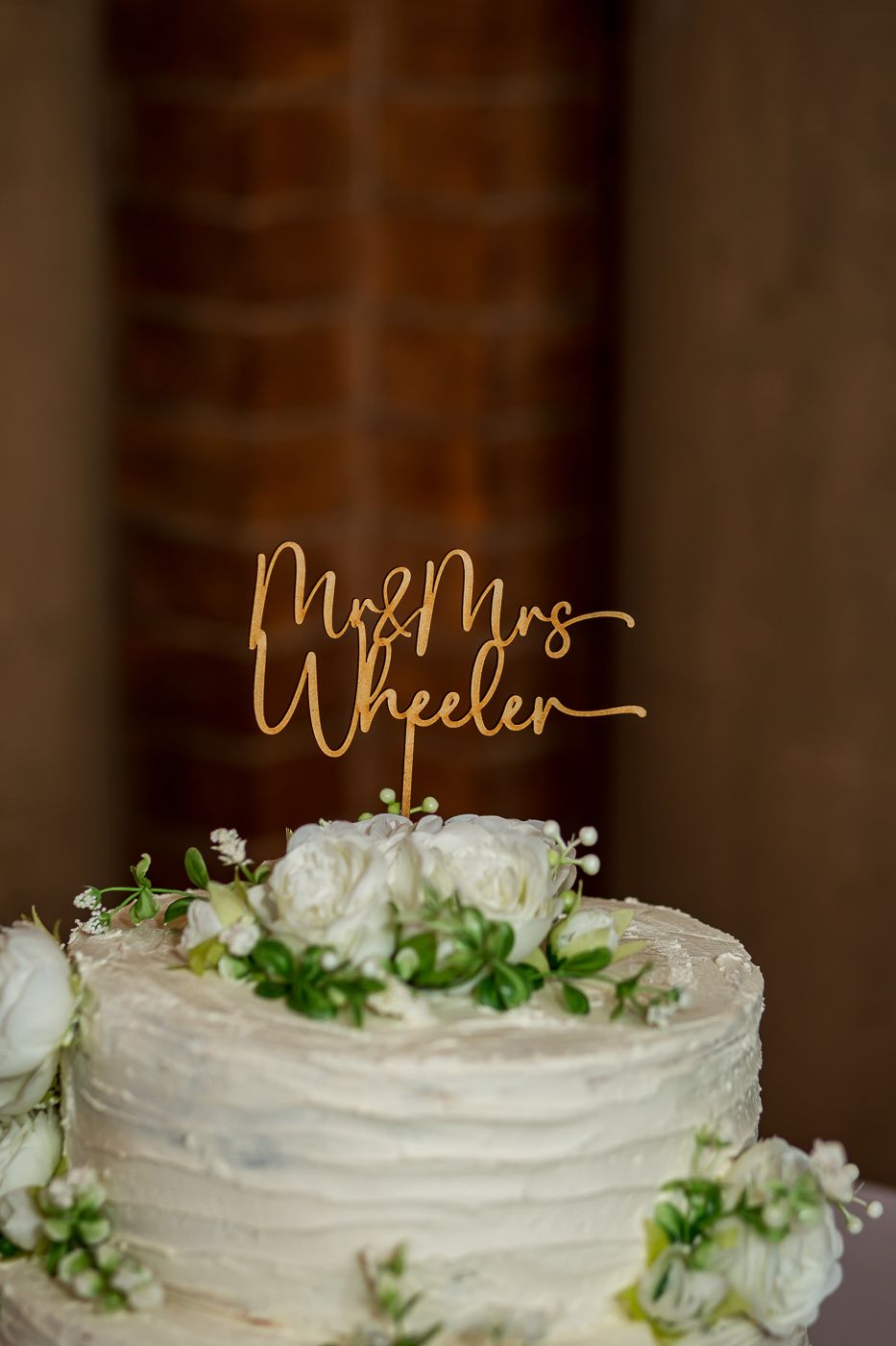 White wedding cake with gold topper and fresh floral decorations on rustic wood slice base.