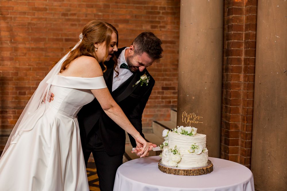 Couple participates in traditional cake cutting ceremony at brick-walled reception venue.