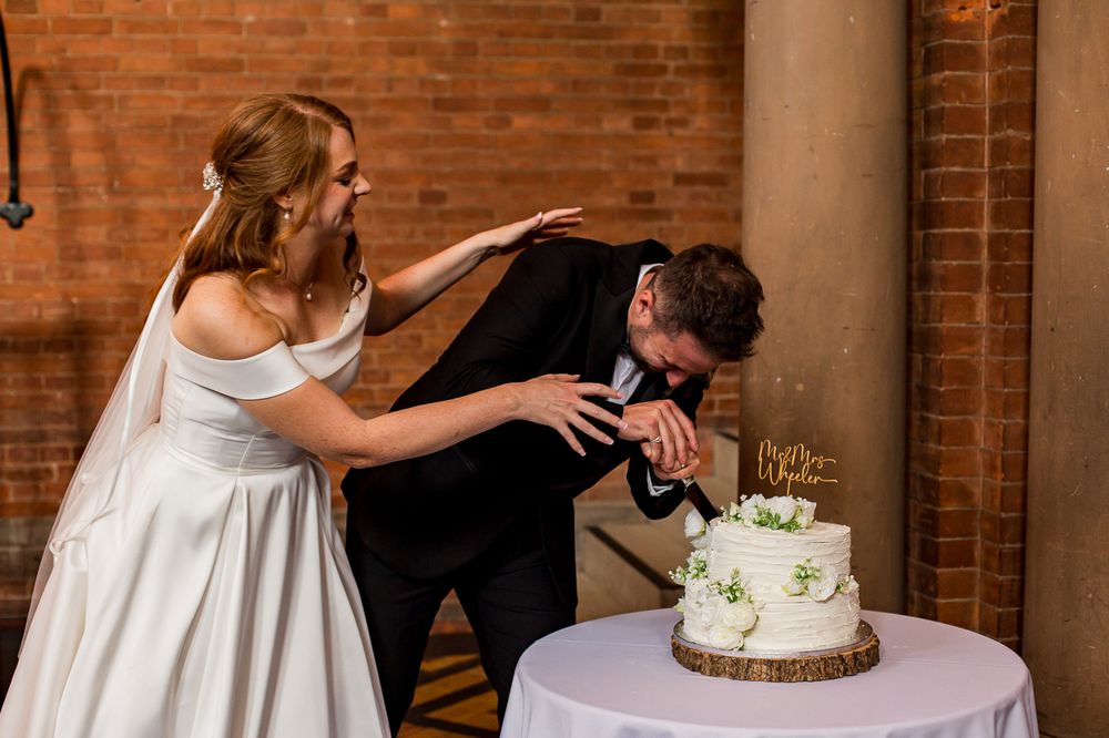 Newlyweds share a playful moment with their wedding cake in a rustic brick venue.