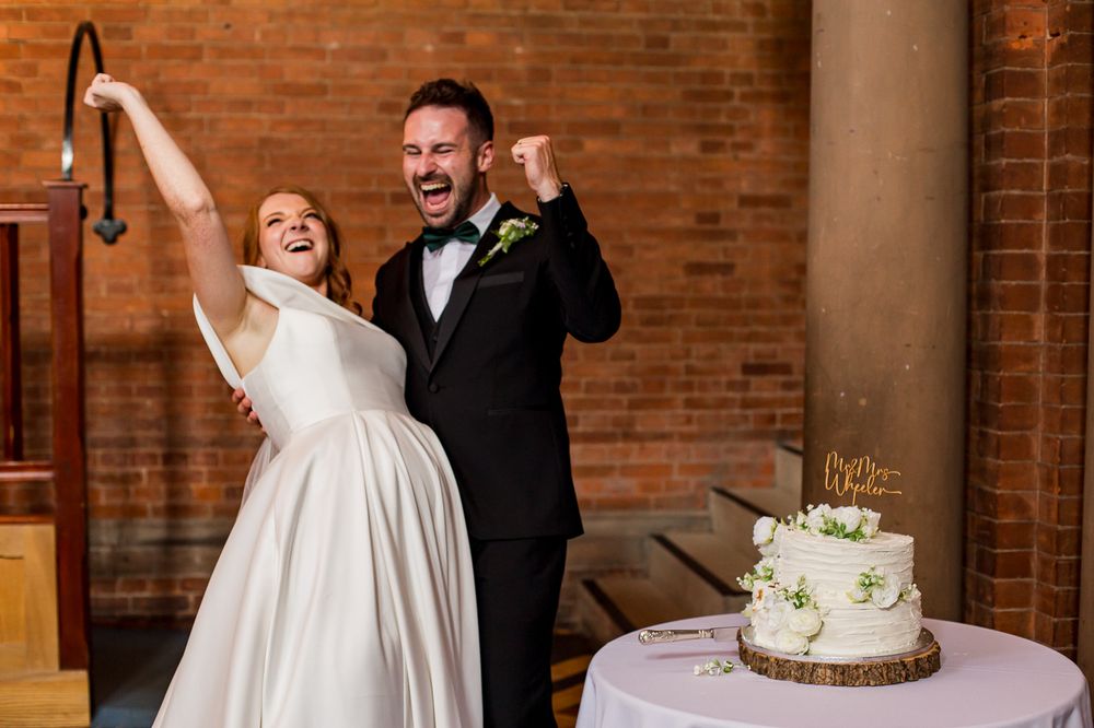 Wedding couple shares joyful moment during cake cutting at reception with brick backdrop.