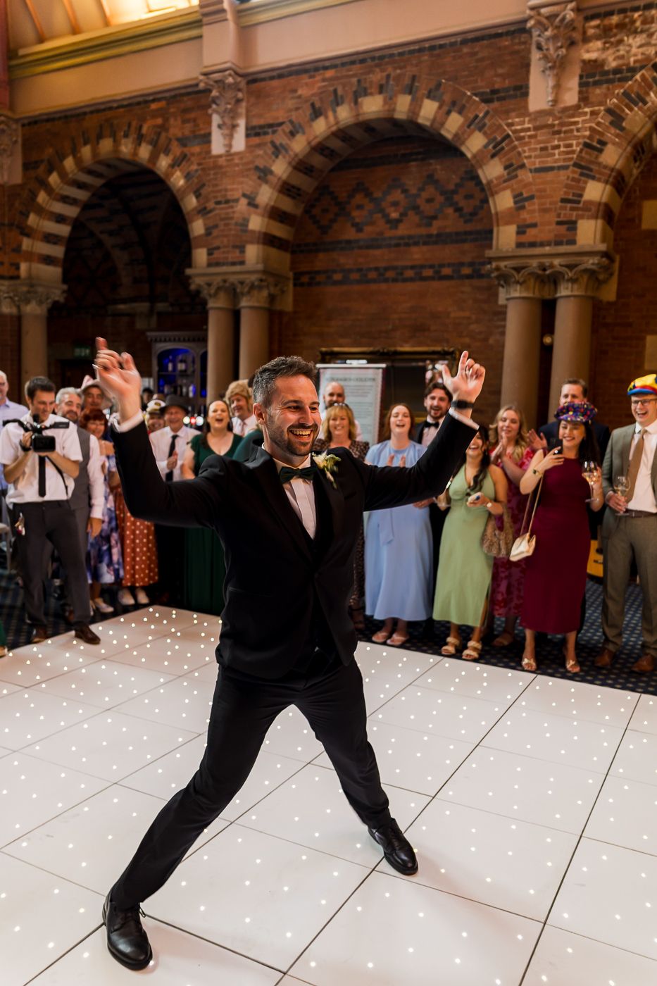 Wedding guests dance on illuminated white dance floor under ornate brick archways.