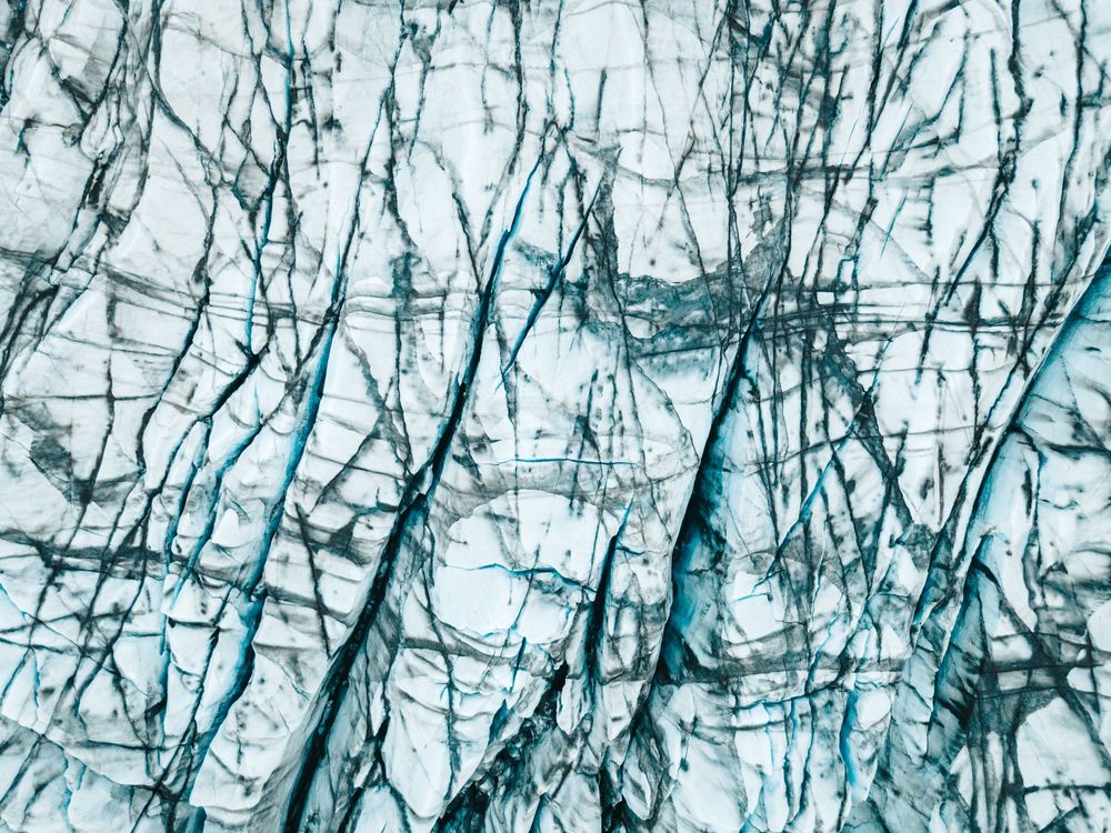 Aerial view of a glacier surface with intricate blue crevasses and cracks forming abstract patterns on white ice.