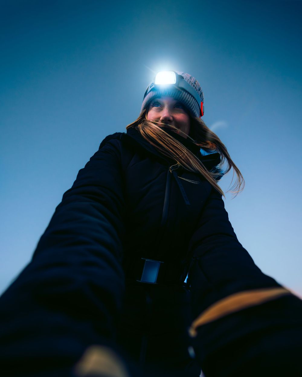 Woman wearing a headlamp and winter hat photographed from below against a clear blue sky.