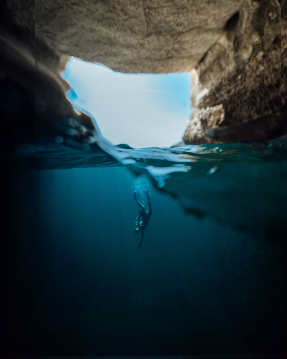 A freediver submerged in crystal-clear blue water inside a sea cave with light streaming through a rocky opening above.