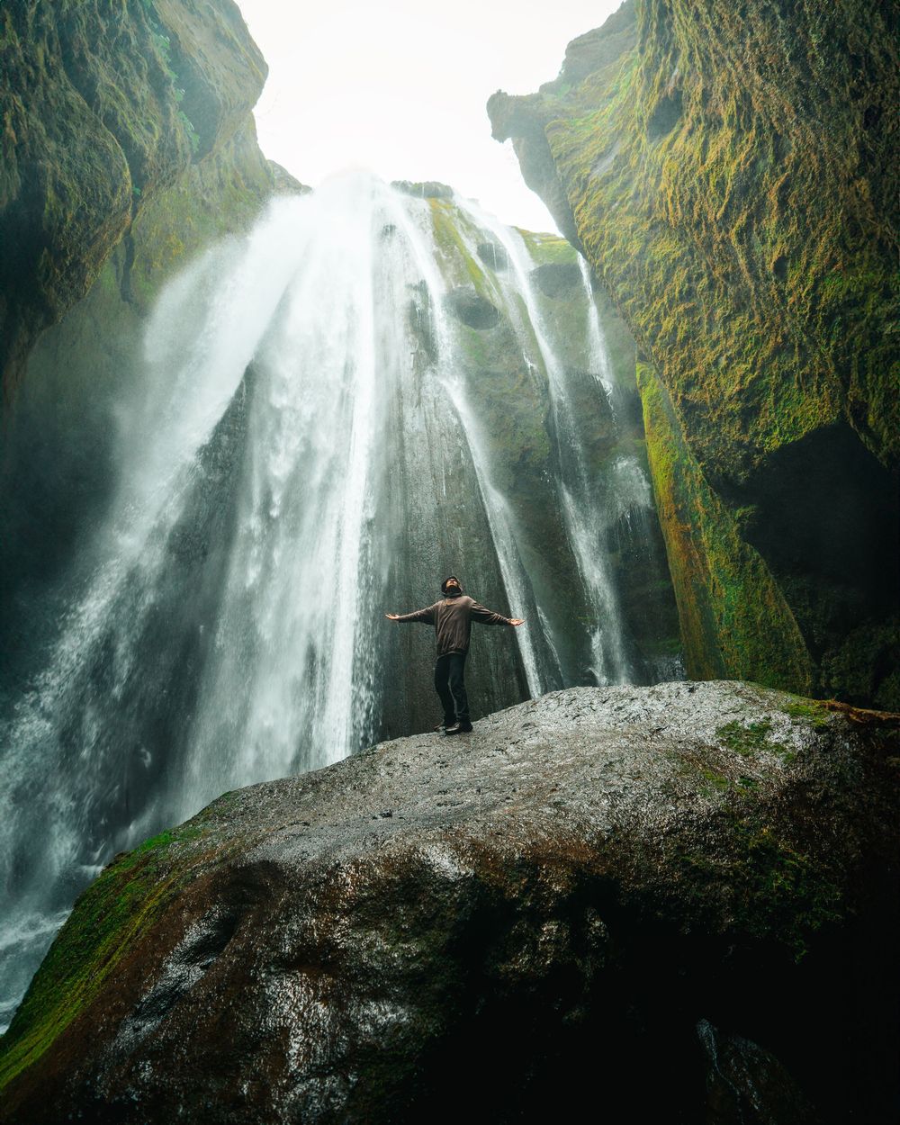A silhouetted figure stands on a mossy rock ledge in front of a majestic cascading waterfall in a lush green gorge.
