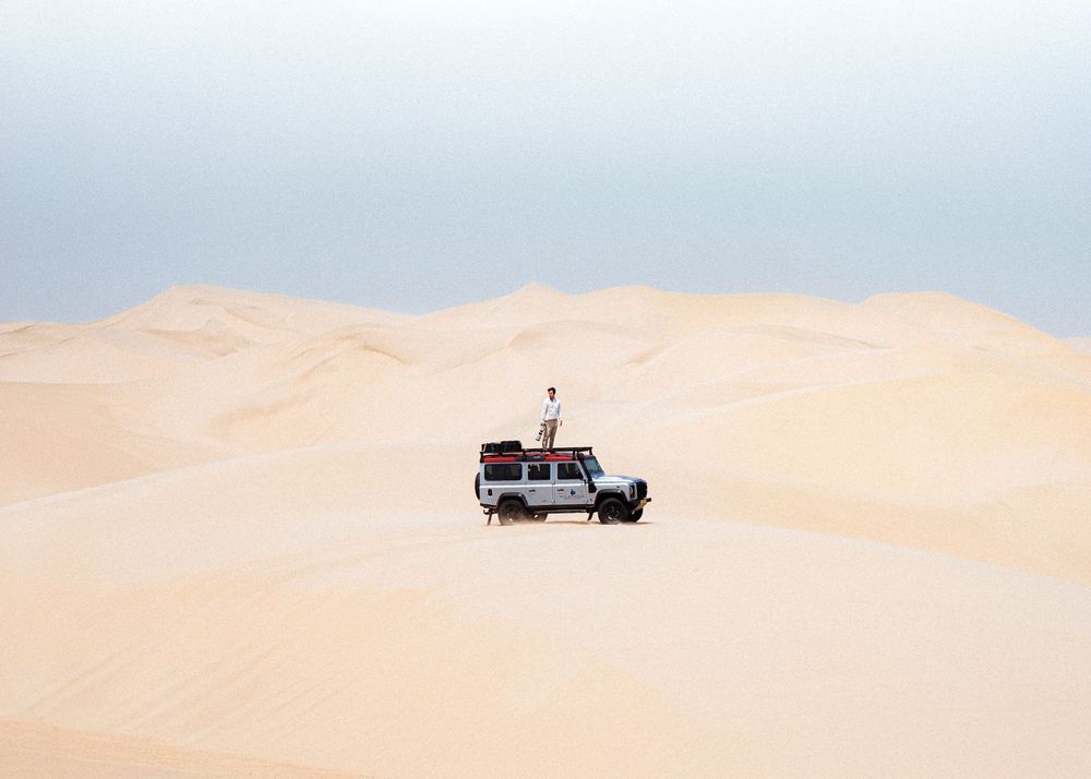 Off-road vehicles traverse vast desert sand dunes under a pale blue sky.