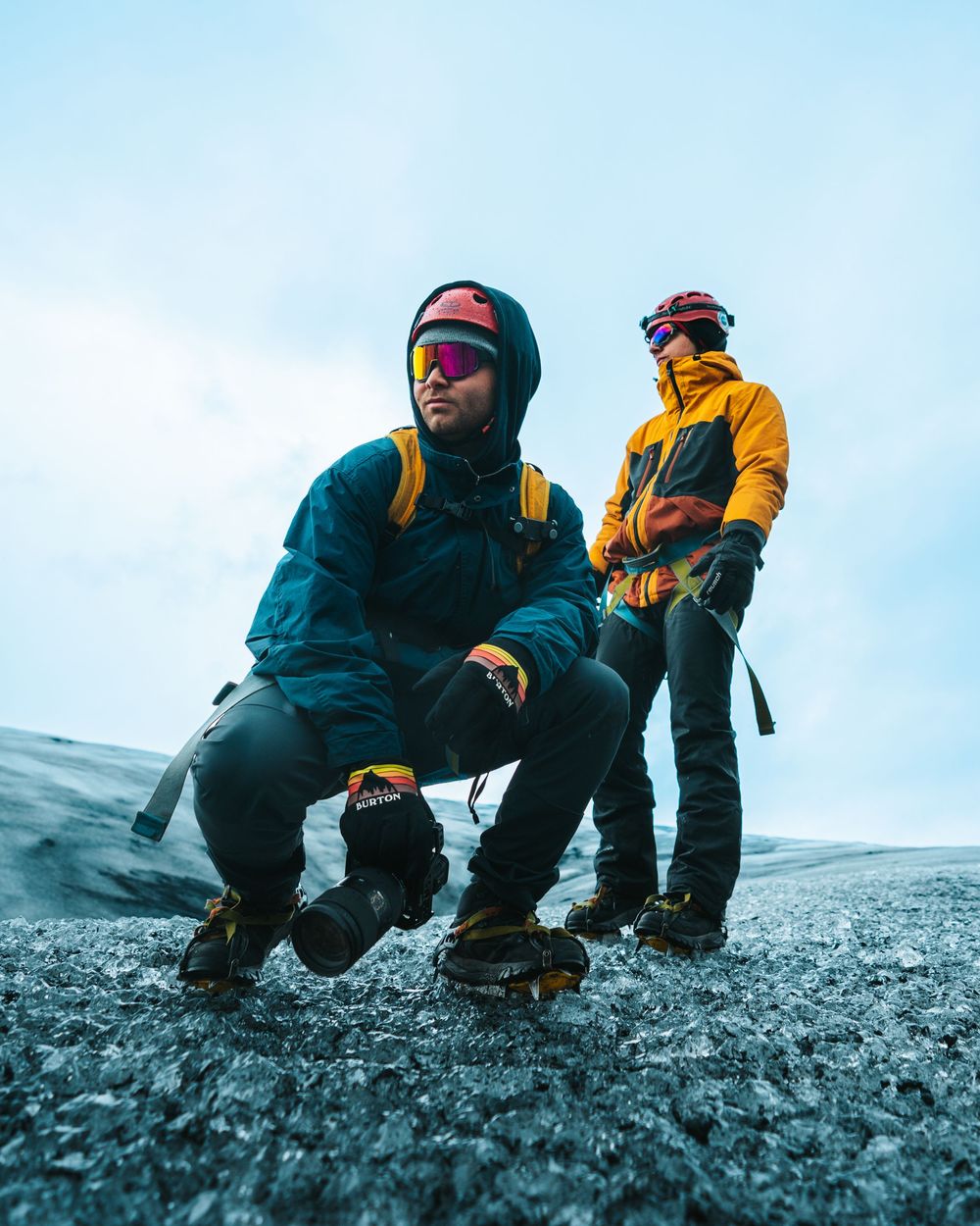Two adventurers in winter gear crouch down to examine the rocky terrain on a cloudy mountainside.