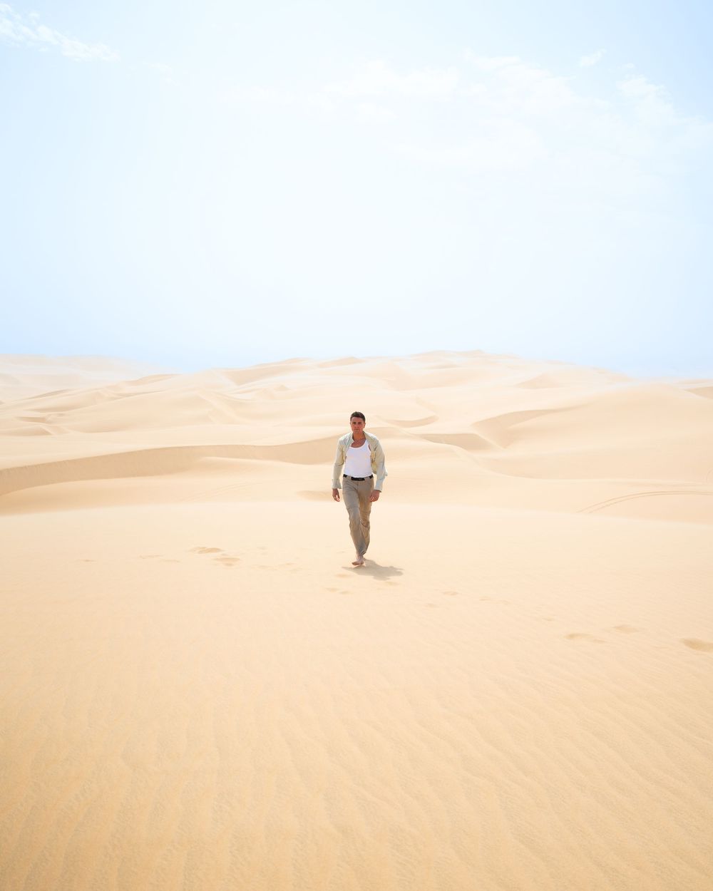 A man walks alone across vast golden sand dunes under a bright hazy sky in a desert landscape.