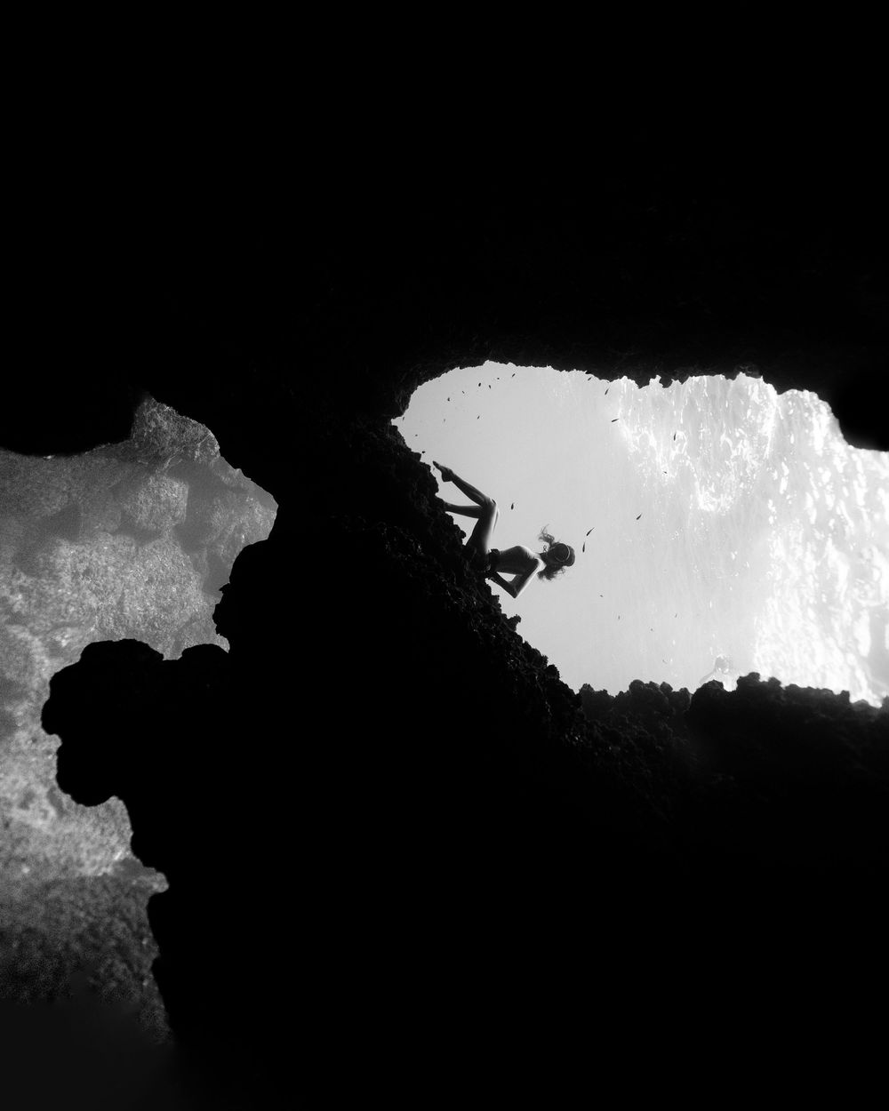 Black and white silhouette of a person climbing through a rocky cave opening with bright light beyond.