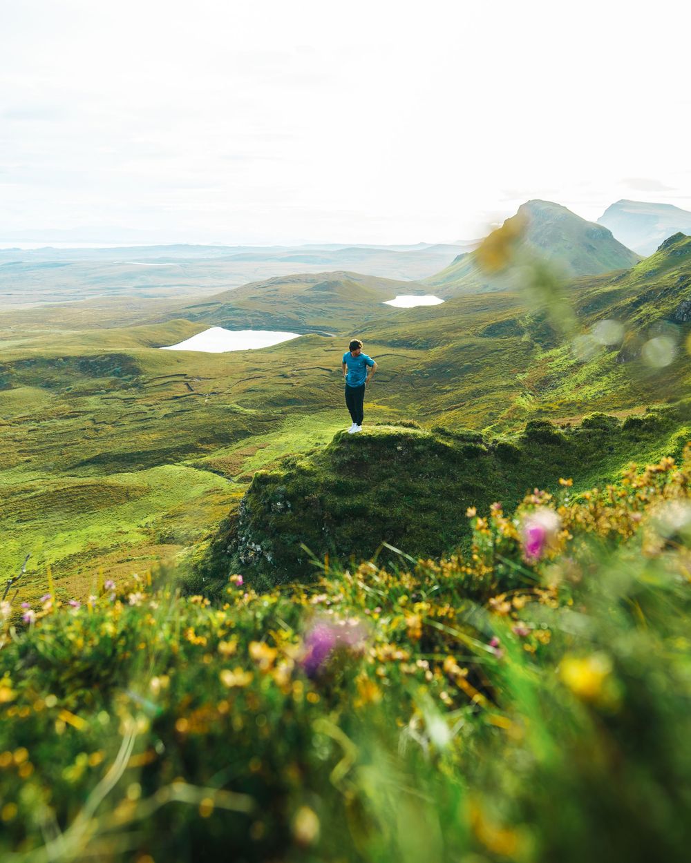 Person standing on a green hilltop overlooking a vast Scottish Highland landscape with lakes and mountains in the distance.