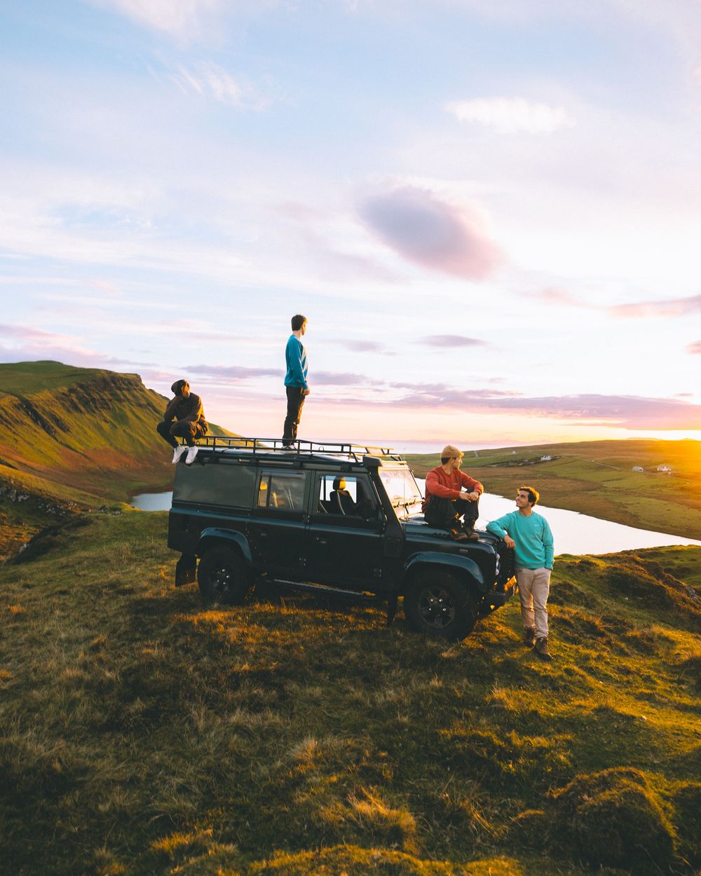Group enjoying sunset on a hillside with Land Rover Defender, standing on vehicle roof against dramatic sky and landscape.