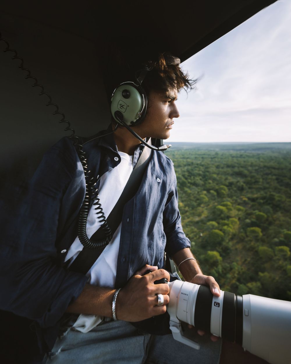 Photographer wearing headphones and holding telephoto lens looks out over green forest canopy from aerial viewpoint.