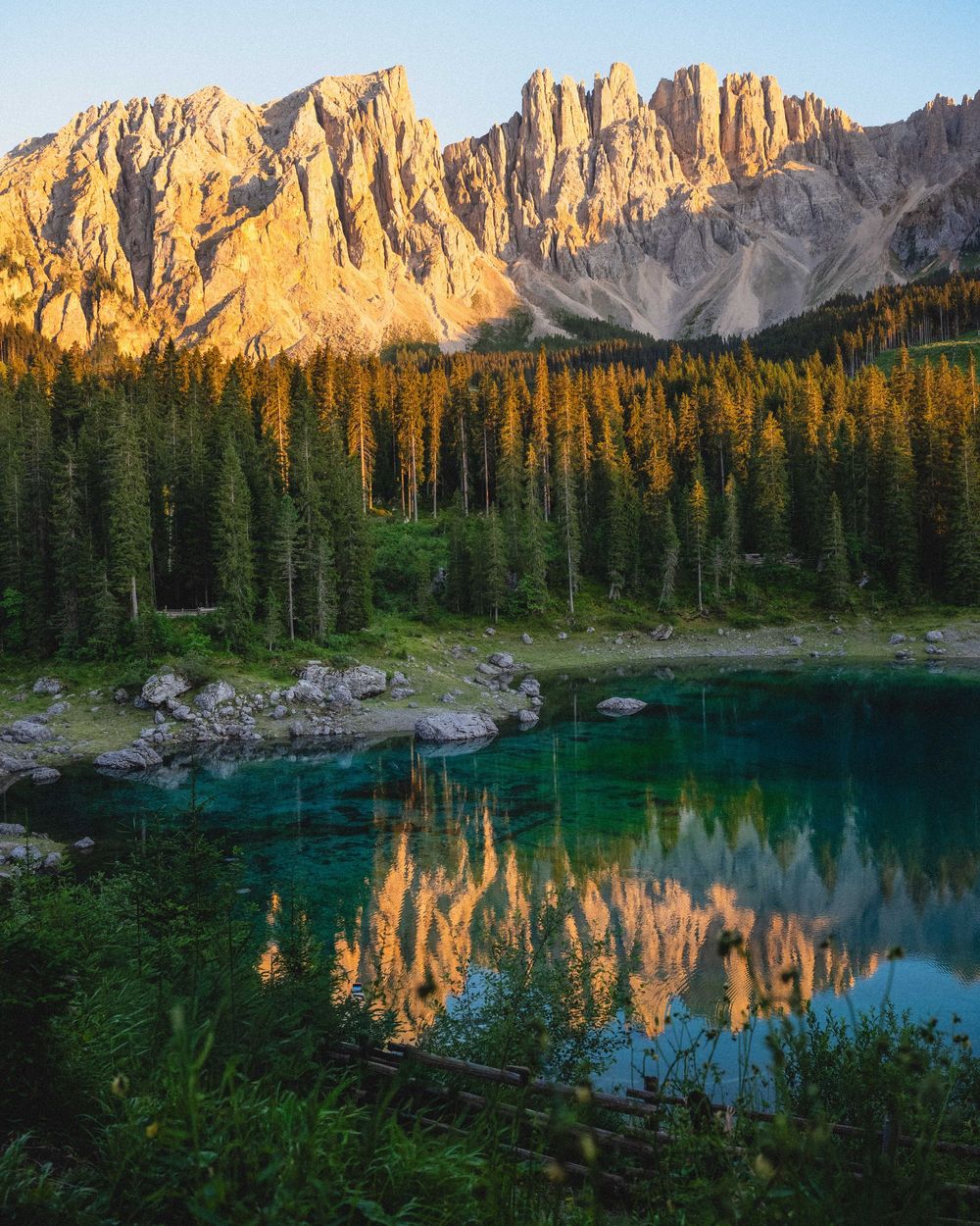 Crystal-clear turquoise alpine lake reflecting golden Dolomite peaks surrounded by lush evergreen forest at sunset.