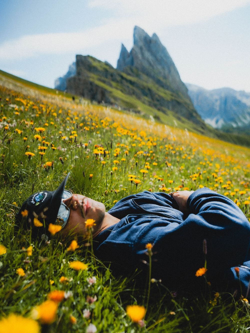 Person lying in a wildflower meadow with sunglasses, relaxing against a backdrop of dramatic mountain peaks.