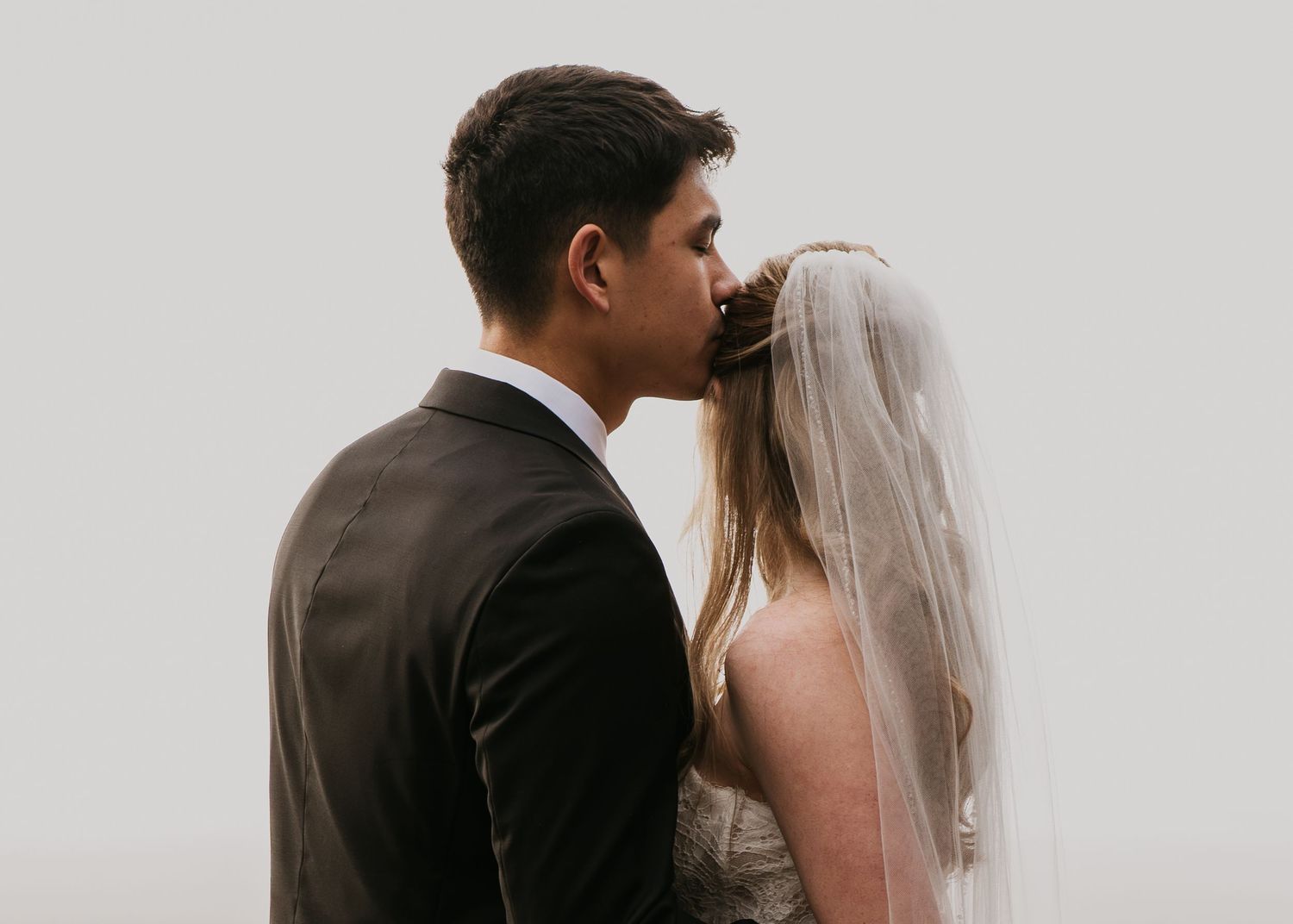 Asian husband kissing Caucasian wife on the wedding day on the side of the head in Seabrook Washington