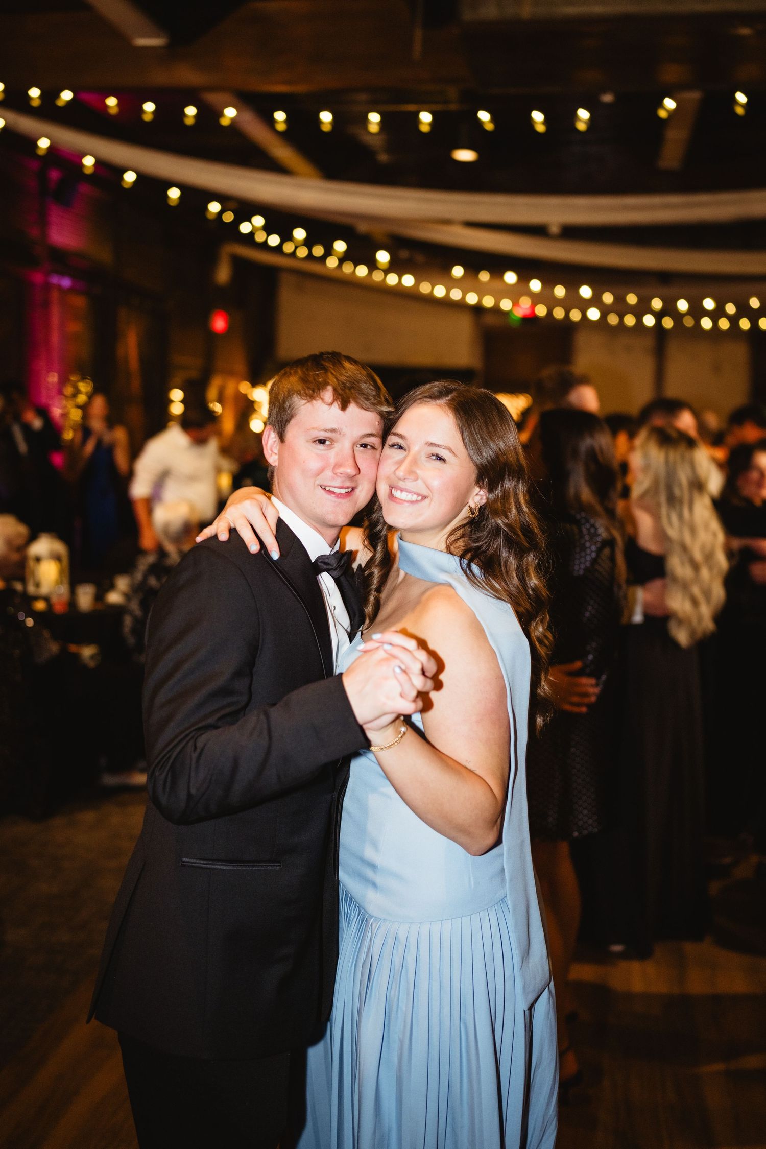 A couple dances together at an indoor reception venue decorated with string lights overhead.