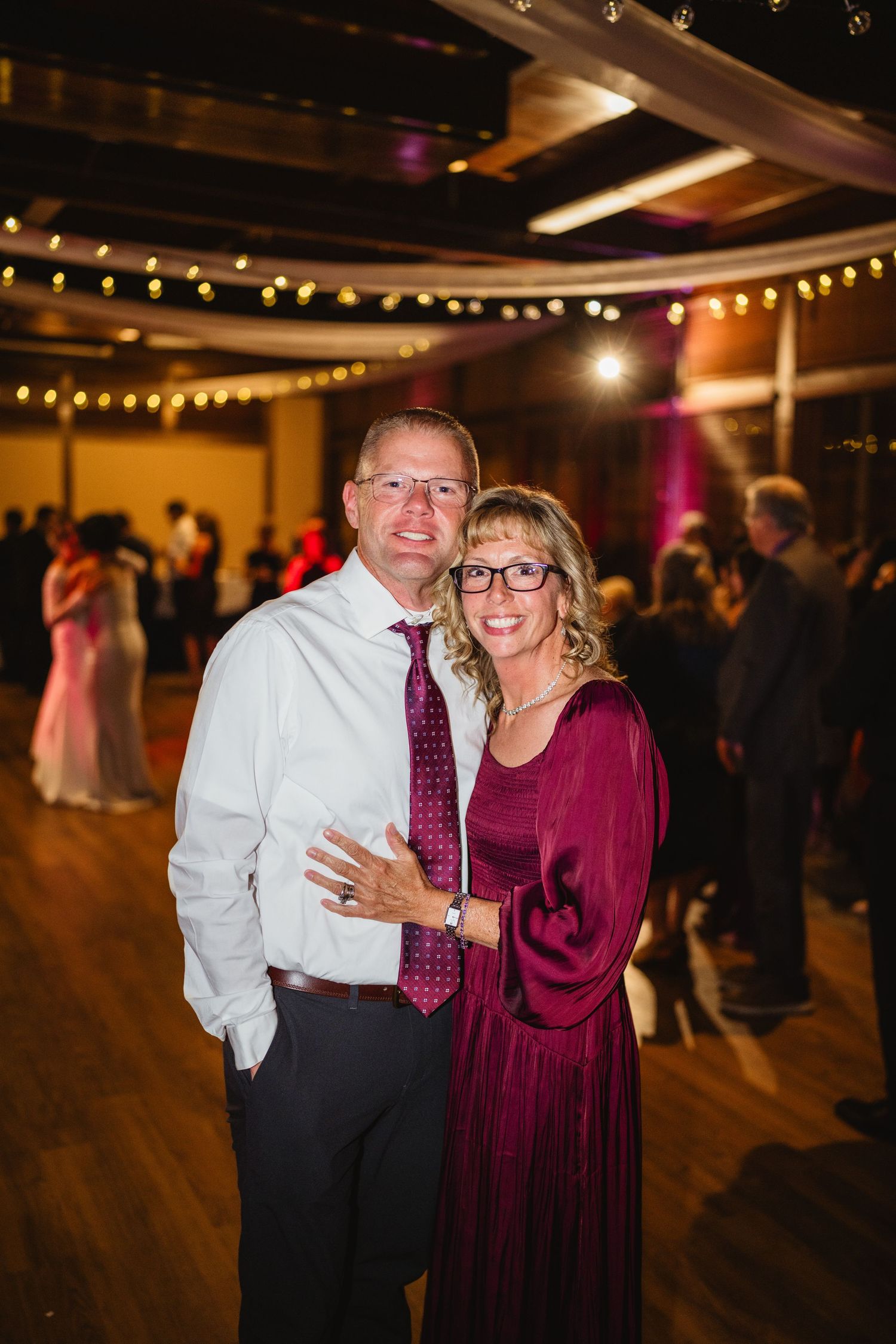 A couple in formal attire dancing at an indoor reception with string lights and wooden floors.