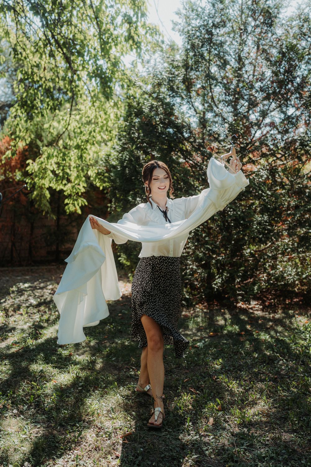 A figure in a flowing white top and patterned skirt dances joyfully in dappled sunlight under trees.
