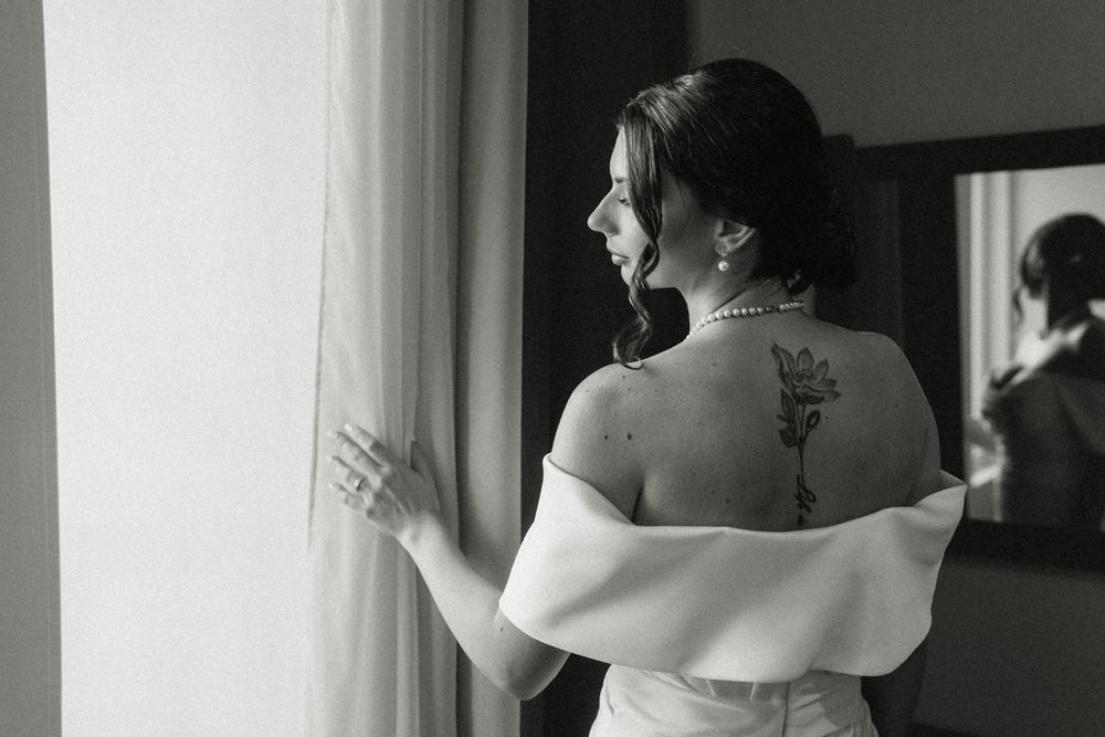 Elegant bride in off-shoulder dress gazes out window, showing delicate back tattoo detail.