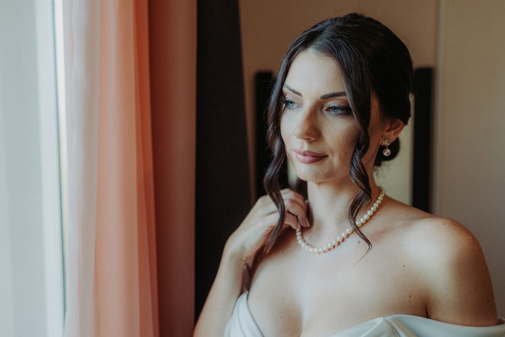 A person in a white strapless dress and pearl necklace gazes softly by a window.