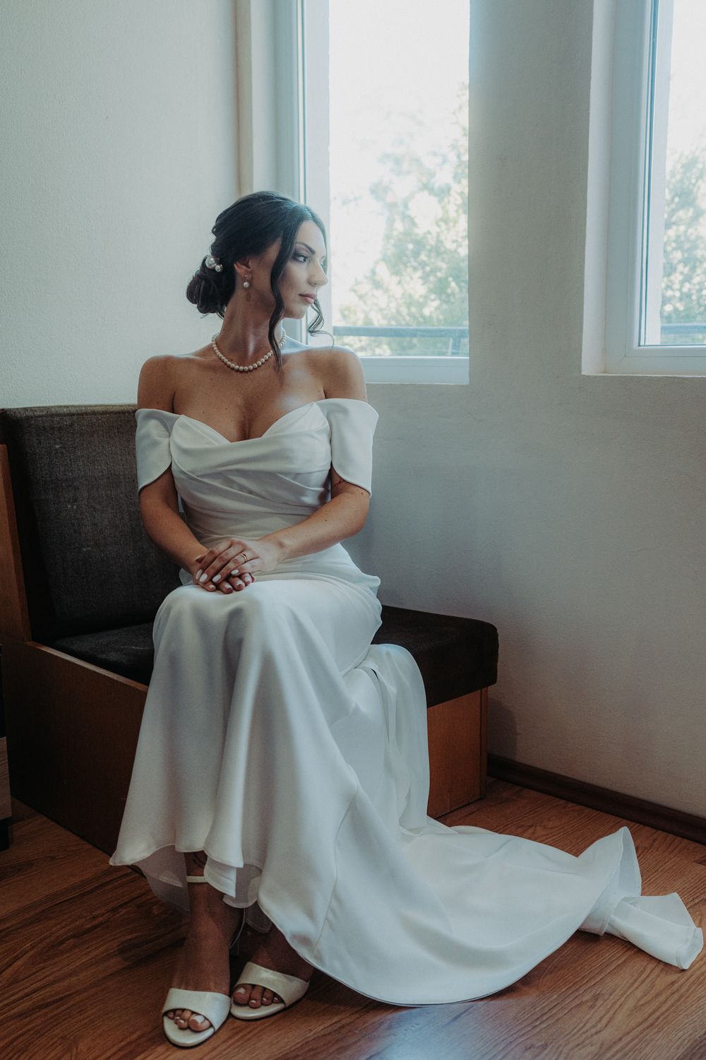 Bride in classic white wedding dress sitting thoughtfully by window in natural light.